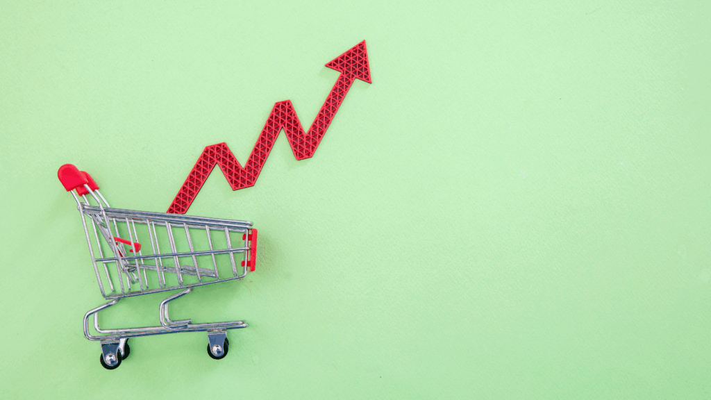 A small shopping cart on a light green background with a red zigzag arrow pointing upward, symbolizing rising prices or increased consumer spending.