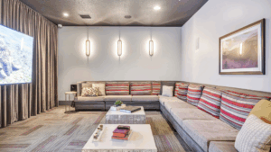 A modern lounge with an L-shaped sofa featuring striped cushions, a large wall-mounted TV, two marble coffee tables, wall art, three vertical lights, and floor-to-ceiling curtains.