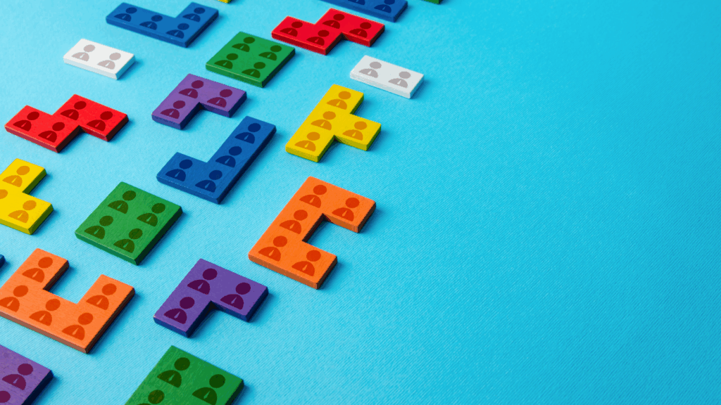 Colorful Tetris-like puzzle pieces with small icons of people are arranged on a blue background, partially scattered and partially forming shapes on the left side of the image. The right side is empty.