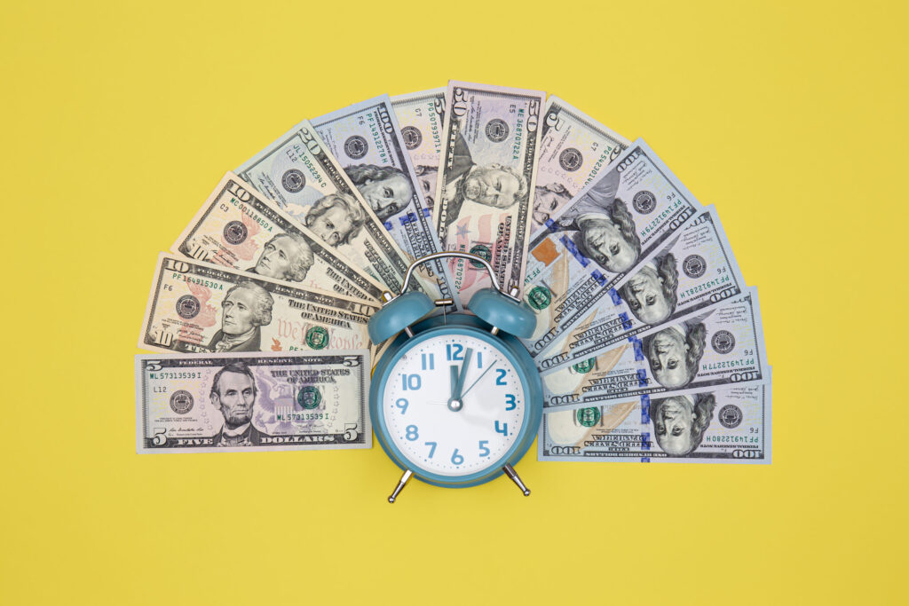 A blue alarm clock sits in front of a fan-shaped arrangement of U.S. dollar bills on a yellow background, symbolizing the concept of time and money.