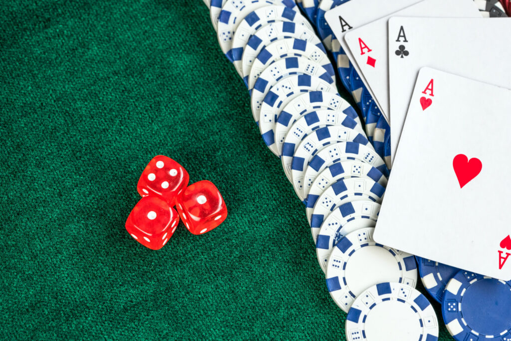 Red dice, blue and white poker chips, and four aces (spades, clubs, hearts, diamonds) are arranged on a green felt casino table.