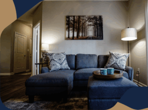 A cozy living room with a dark blue sectional sofa, patterned pillows, a floor lamp, a wall art of a forest, and a wooden tray with mugs on an ottoman. Warm lighting and neutral walls create a relaxing atmosphere.
