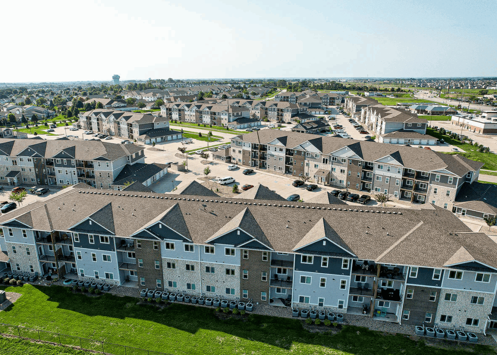 Aerial view of a large apartment complex with multiple three-story buildings, parking lots, and green lawns, surrounded by suburban neighborhoods under a clear sky.