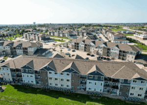 Aerial view of a large apartment complex with multiple three-story buildings, parking lots, and green lawns, surrounded by suburban neighborhoods under a clear sky.