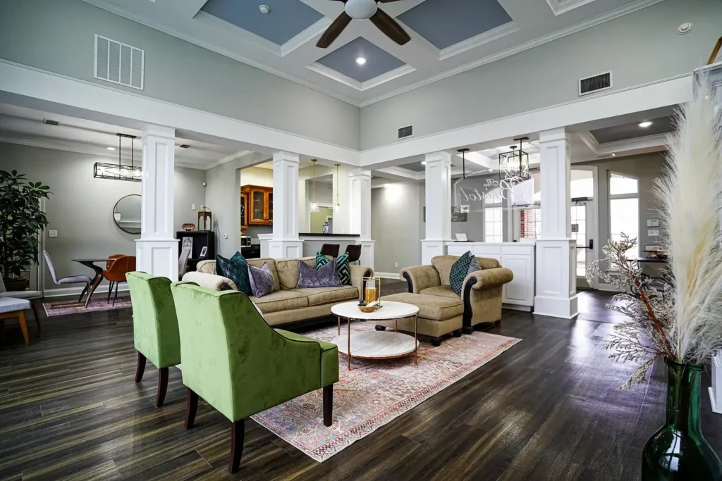 A modern, spacious living room with high ceilings, green armchairs, a beige sofa, a round coffee table, and large windows letting in natural light. Decorative plants and elegant columns accent the open-concept space.