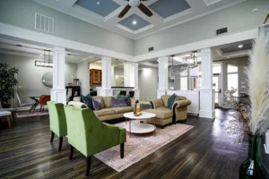 A modern, spacious living room with high ceilings, green armchairs, a beige sofa, a round coffee table, and large windows letting in natural light. Decorative plants and elegant columns accent the open-concept space.