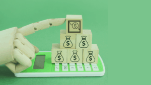 A wooden robotic hand points at wooden blocks stacked on a green calculator. The blocks display money bag icons and a chart with a dollar sign, symbolizing finance and technology. The background is solid green.