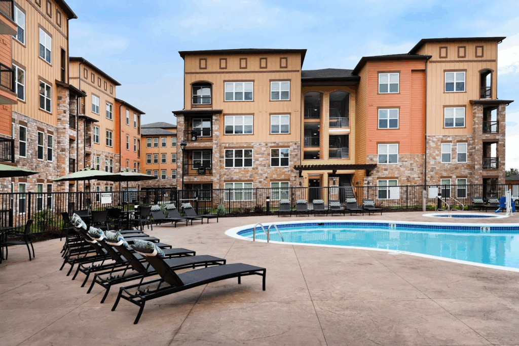 Modern apartment complex with three- and four-story buildings, featuring balconies, stone and orange siding, and a fenced outdoor pool area surrounded by lounge chairs and umbrellas on a spacious patio.