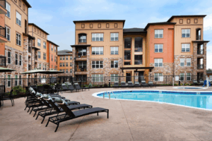 Modern apartment complex with three- and four-story buildings, featuring balconies, stone and orange siding, and a fenced outdoor pool area surrounded by lounge chairs and umbrellas on a spacious patio.