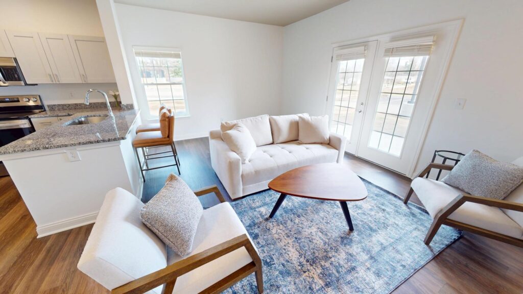 A bright living room with a white sofa, two cushioned chairs, a wooden coffee table on a blue rug, and large windows with glass doors. The kitchen with bar stools and granite counters is visible to the left.