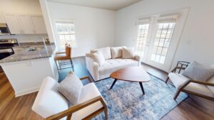 A bright living room with a white sofa, two cushioned chairs, a wooden coffee table on a blue rug, and large windows with glass doors. The kitchen with bar stools and granite counters is visible to the left.