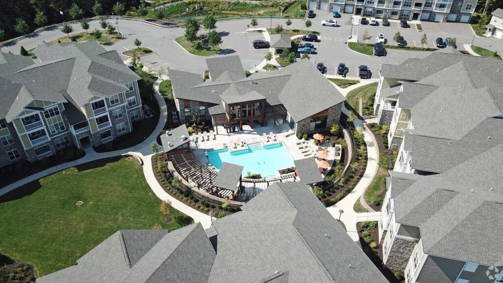 Aerial view of an apartment complex featuring a central swimming pool, poolside lounge chairs, umbrellas, landscaped pathways, and surrounding gray-roofed buildings with parked cars nearby.