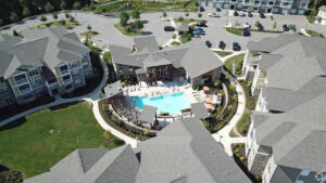 Aerial view of an apartment complex featuring a central swimming pool, poolside lounge chairs, umbrellas, landscaped pathways, and surrounding gray-roofed buildings with parked cars nearby.
