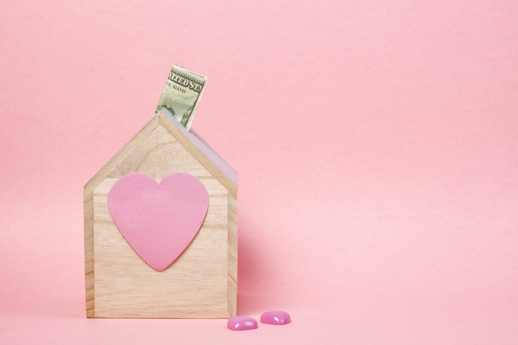A wooden house-shaped box with a pink heart on the front holds a dollar bill. Two small pink heart-shaped objects lie on the pink surface in front of the box, against a pink background.