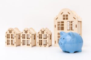 A blue piggy bank sits in front of four wooden model houses of different sizes, all placed against a plain white background.