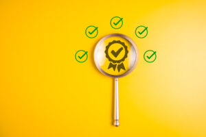 A magnifying glass focuses on an award ribbon with a check mark inside, surrounded by five green check marks on a bright yellow background, symbolizing quality assurance or approval.