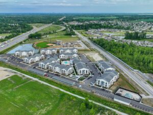 Aerial view of a modern apartment complex with multiple white buildings, parking lots, a swimming pool, nearby roads, a baseball field, and surrounding green areas and residential neighborhoods.