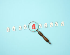 A magnifying glass focuses on a red house icon among several white house icons arranged in a row on a light blue background.
