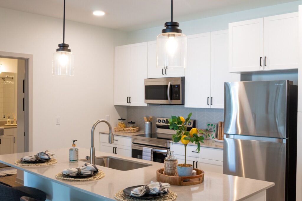Modern kitchen with white cabinets, stainless steel appliances, and a center island set with plates and napkins. Pendant lights hang above the island, which also features a sink and decorative tray with plants and lemons.