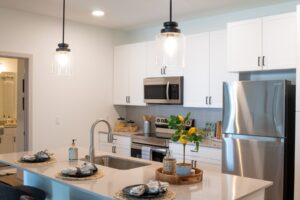 Modern kitchen with white cabinets, stainless steel appliances, and a center island set with plates and napkins. Pendant lights hang above the island, which also features a sink and decorative tray with plants and lemons.