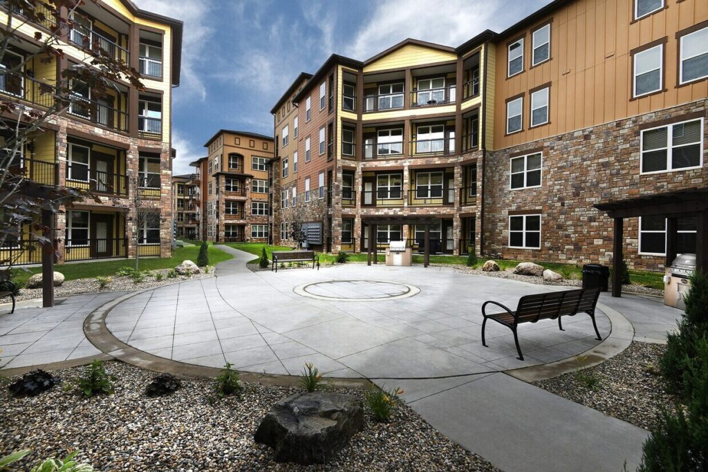 A modern apartment complex features multi-story buildings with balconies, stone and wood exteriors, and a landscaped courtyard with benches, circular paved walkway, rocks, and small plants under a partly cloudy sky.
