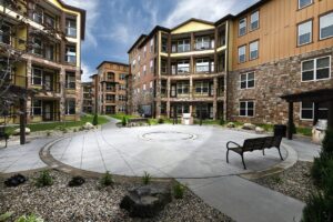 A modern apartment complex features multi-story buildings with balconies, stone and wood exteriors, and a landscaped courtyard with benches, circular paved walkway, rocks, and small plants under a partly cloudy sky.
