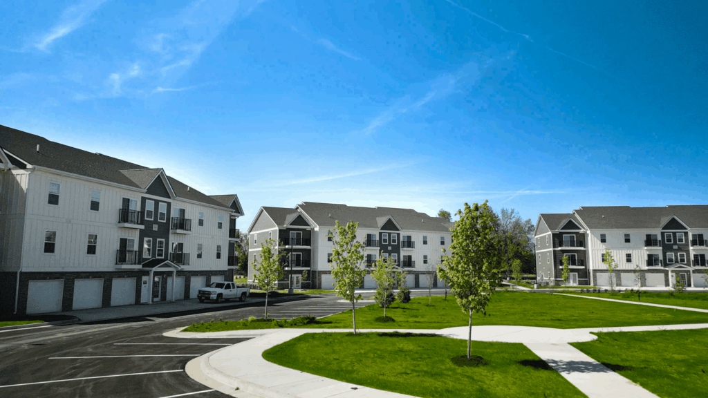 A row of modern, three-story apartment buildings with garages sits beside a green lawn and young trees under a clear blue sky in a residential neighborhood.