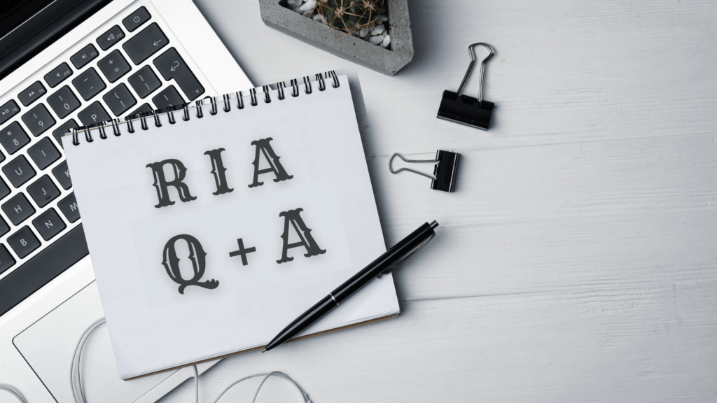 A spiral notebook on a desk displays the text RIA Q + A. Nearby are a laptop keyboard, pen, binder clips, earphones, and a small potted plant.