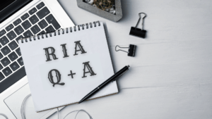 A spiral notebook on a desk displays the text RIA Q + A. Nearby are a laptop keyboard, pen, binder clips, earphones, and a small potted plant.