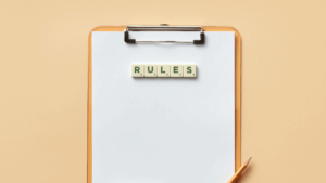 A blank clipboard with the word RULES spelled out in letter tiles at the top, and an orange pencil positioned on the lower right corner, set against a beige background.