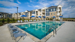 A clear outdoor swimming pool with lounge chairs, small tables, and blue umbrellas sits in front of modern, multi-story apartment buildings under a partly cloudy sky.