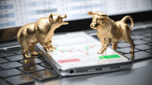 Two golden figurines of a bear and a bull stand on a smartphone displaying a stock chart, placed on a laptop keyboard with financial data visible on the computer screen in the background.