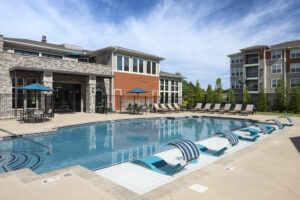 A modern outdoor swimming pool with poolside loungers, umbrella-covered tables, and a large apartment building in the background on a sunny day.