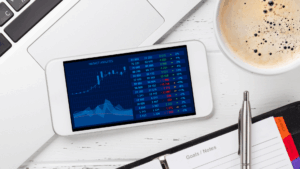 A smartphone displays a financial market analysis with charts and data, placed on a desk next to a laptop, a cup of coffee, a pen, and a notebook labeled Goals / Notes.