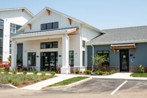A modern building with white and blue-gray siding, large windows, a covered entrance, visible house numbers, landscaped shrubs, and an empty parking lot in front under a clear sky.