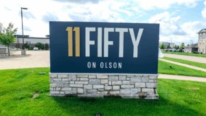 Large outdoor sign reading 11 FIFTY ON OLSON with gold and white text on a blue background, mounted on a stone base, on a grassy lawn near a parking lot and buildings.
