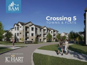 People walk on a curved sidewalk in front of modern, three-story apartment buildings under a clear blue sky. Signs read Crossing 5 Towns & Flats, with BAM Capital and JC Hart logos visible.