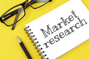 A notebook with the words Market research written on it, placed on a yellow background next to a pair of black glasses and a pen.