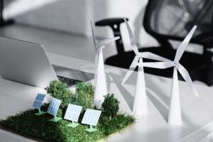 A desk with a laptop, two model wind turbines, and a miniature landscape featuring solar panels and trees, representing renewable energy and sustainability concepts.