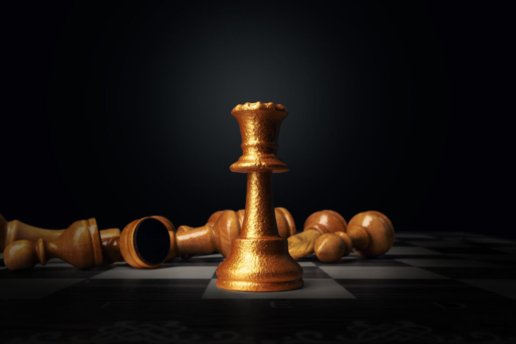 A golden chess queen stands upright on a chessboard, illuminated in the center, while several wooden chess pieces lie toppled in the background against a dark backdrop.