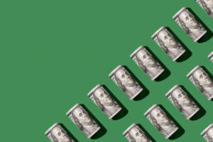 Rolled-up US hundred-dollar bills arranged in a diagonal, upward trend pattern on a solid green background, casting shadows to the right.