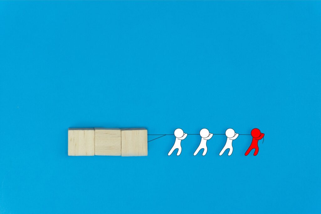 Four illustrated figures, three white and one red, are pulling a string attached to three wooden blocks against a blue background, symbolizing teamwork and leadership.