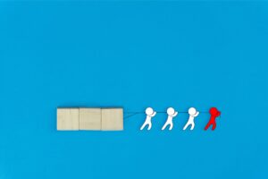 Four illustrated figures, three white and one red, are pulling a string attached to three wooden blocks against a blue background, symbolizing teamwork and leadership.