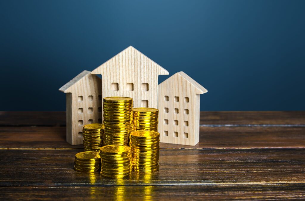 Stacks of gold coins are arranged in front of three small wooden house models on a wooden table, with a blue background.