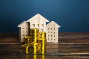 Stacks of gold coins are arranged in front of three small wooden house models on a wooden table, with a blue background.