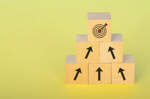 Four wooden blocks are stacked in a pyramid on a yellow background. Three lower blocks have black upward arrows, and the top block shows a target with an arrow hitting the center.