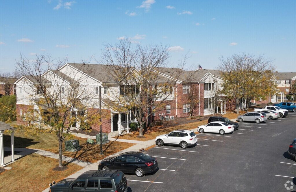 A row of two-story brick and white apartment buildings with leafless trees in front, adjacent to a parking lot filled with cars under a clear blue sky.