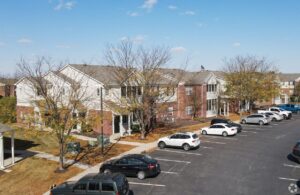 A row of two-story brick and white apartment buildings with leafless trees in front, adjacent to a parking lot filled with cars under a clear blue sky.