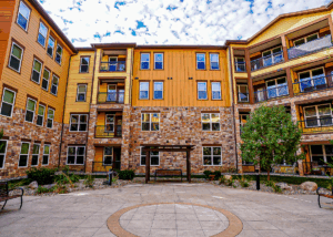 A three-story apartment building with yellow and brown siding and stone accents, featuring balconies and windows, surrounds a courtyard with a circular design on the ground and landscaping with bushes and trees.