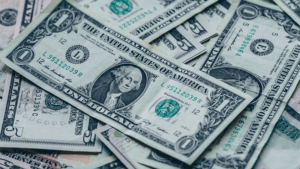 A close-up view of several scattered U.S. one-dollar bills, showing the front side with George Washington’s portrait and various serial numbers and seals visible.